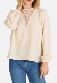 Blusa Beige con inserto de encaje en el cuello, mangas largas abullonadas con puños fruncidos y un corte relajado, combinada con jeans azules.
