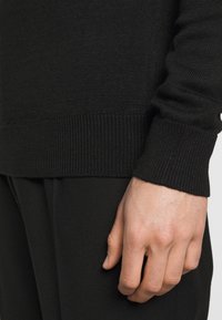 Czarny sweter z dzianiny, z detalami takimi jak prążkowane mankiety i dół, o gładkiej teksturze. Ręka spoczywa na ciemnej tkaninie poniżej, co podkreśla kontrast kolorów.