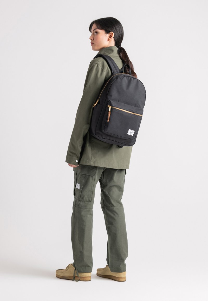 Mochila negra con cremalleras doradas, forma rectangular y un pequeño bolsillo frontal. El modelo lleva puesto una chaqueta y pantalones verdes, con zapatos marrones.