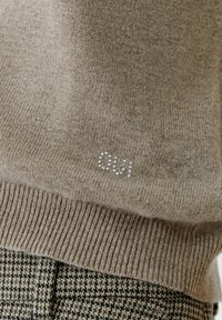 Beige gestrickter Pullover mit geripptem Saum, der auf der Vorderseite "Oui" in schimmernden Perlen trägt. Kombiniert mit schwarz-weißen Hahnentritt-Hosen.