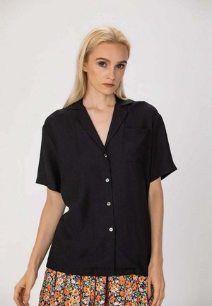 Femme blonde portant une chemise noire ample à manches courtes et une jupe fleurie, debout devant un fond clair uni.