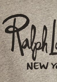 Texte en lettres cursives noires "Ralph L" et lettres majuscules "NEW Y" imprimés sur un tissu gris, probablement une partie d'une phrase imprimée plus longue ou d'un nom de marque.