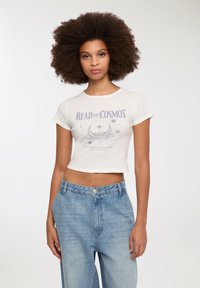 Biała krótka koszulka z grafiką "READ the COSMOS" w kolorze fioletowym. Połączona z jasnoniebieskimi, wysokim stanem dżinsami. Gładka faktura.