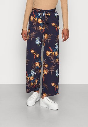 Granatowe spodnie o szerokich nogawkach z pomarańczowym, niebieskim i białym kwiatowym nadrukiem, ze związanym w talii pasem, zestawione z białymi sneakersami na platformie i beżowym prążkowanym topem z długim rękawem.