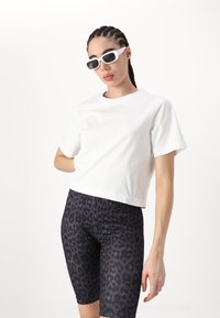 T-shirt blanc court en coton avec des manches courtes, associé à un short de cyclisme noir à imprimé léopard. Les accessoires comprennent des lunettes de soleil rectangulaires blanches.