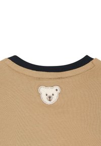 Camisa de algodón color marrón claro con un cuello negro y un parche bordado de la cara de un oso blanco en la parte de atrás. Textura suave, diseño simple.