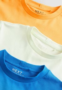 Trois t-shirts à col rond pliés en orange, blanc et bleu, chacun étiqueté avec le nom de la marque « NEXT » à l'intérieur du col.