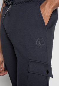 Donker marineblauwe cargobroek van zacht materiaal, met een trekkoord in de taille, zijzakken en een ronde logo-opdruk op de dij.