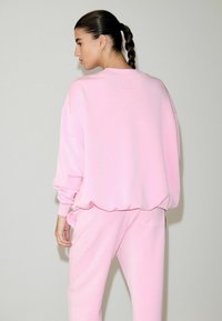Ružová mikina a zodpovedajúce nohavice, vyrobené z mäkkej látky. Mikina má znížené ramená a rebrované manžety, s minimálnym logom ako detailom.