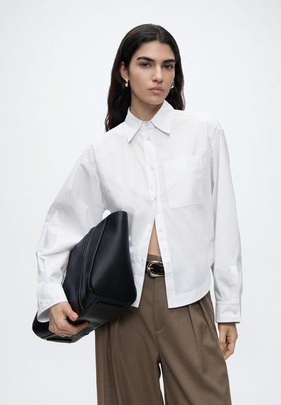 Femme aux longs cheveux foncés portant une chemise blanche boutonnée et un pantalon marron, tenant un grand sac en cuir noir sous son bras.