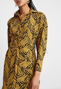 Frau trägt ein langärmliges, tailliertes Kleid mit Senfgelb und schwarzem Zebramuster, mit einem Kragen und goldenen Creolen.