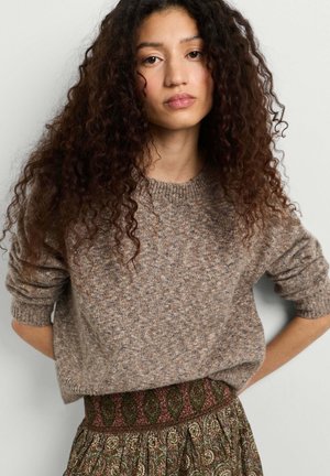 Donna con lunghi capelli ricci che indossa un maglione marrone lavorato a maglia e una gonna verde a fantasia, in piedi davanti a uno sfondo chiaro e uniforme.