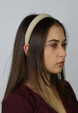 Beżowa, teksturowana opaska wykonana z miękkiego materiału, spoczywająca na prostych, ciemnych włosach. Widok z profilu podkreśla kształt i wzór dodatku.