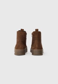 Botas de tobillo de ante marrón con un diseño texturizado, cremalleras laterales y lengüetas de tirón, que cuentan con una suela de goma gruesa y detalles de costura a lo largo del borde.
