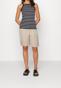 Femme portant un haut sans manches rayé noir et blanc, un short beige montant jusqu'au genou et des sandales ouvertes à bout noir à enfiler, se tenant devant un mur blanc.