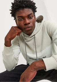 Sudadera gris clara con un bolsillo frontal, cordones y puños acanalados. Presenta una banda de contraste más oscura y un logotipo bordado en el pecho.