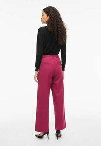 Pull noir ajusté associé à un pantalon taille haute à jambes larges de couleur rose foncé, doté d'une ceinture élastique et de poches arrière, porté avec des talons noirs.