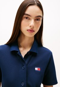 Jūras zilas polo krekliņš ar strukturētu audumu, pogu plati, un mazu izšūtu logo, kas sastāv no sarkanās, baltās un zilās krāsas uz krūšu daļas.