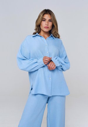 Femeie purtând o cămașă lejeră bleu deschis cu nasturi și pantaloni largi asortați, ajustând manșeta cu o expresie neutră pe fundal gri.