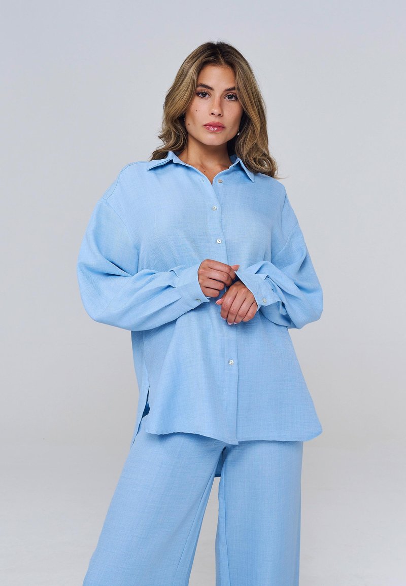 Femme portant une chemise bleu clair ample à boutons et un pantalon large assorti, ajustant le poignet avec une expression neutre sur fond gris.