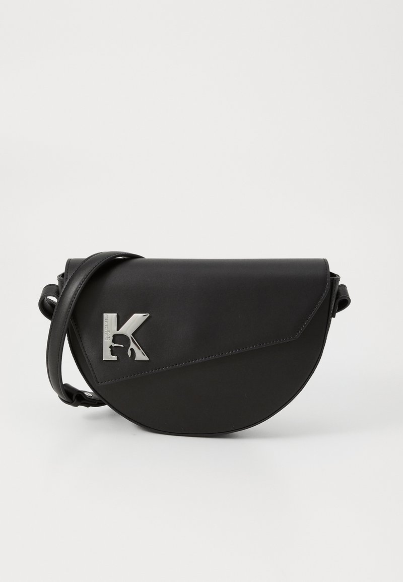 Svart lädercrossbody-väskan har en rundad form, en silverfärgad "K"-logotyp, en sned flap och en smal justerbar rem. Slät textur med minimala detaljer.