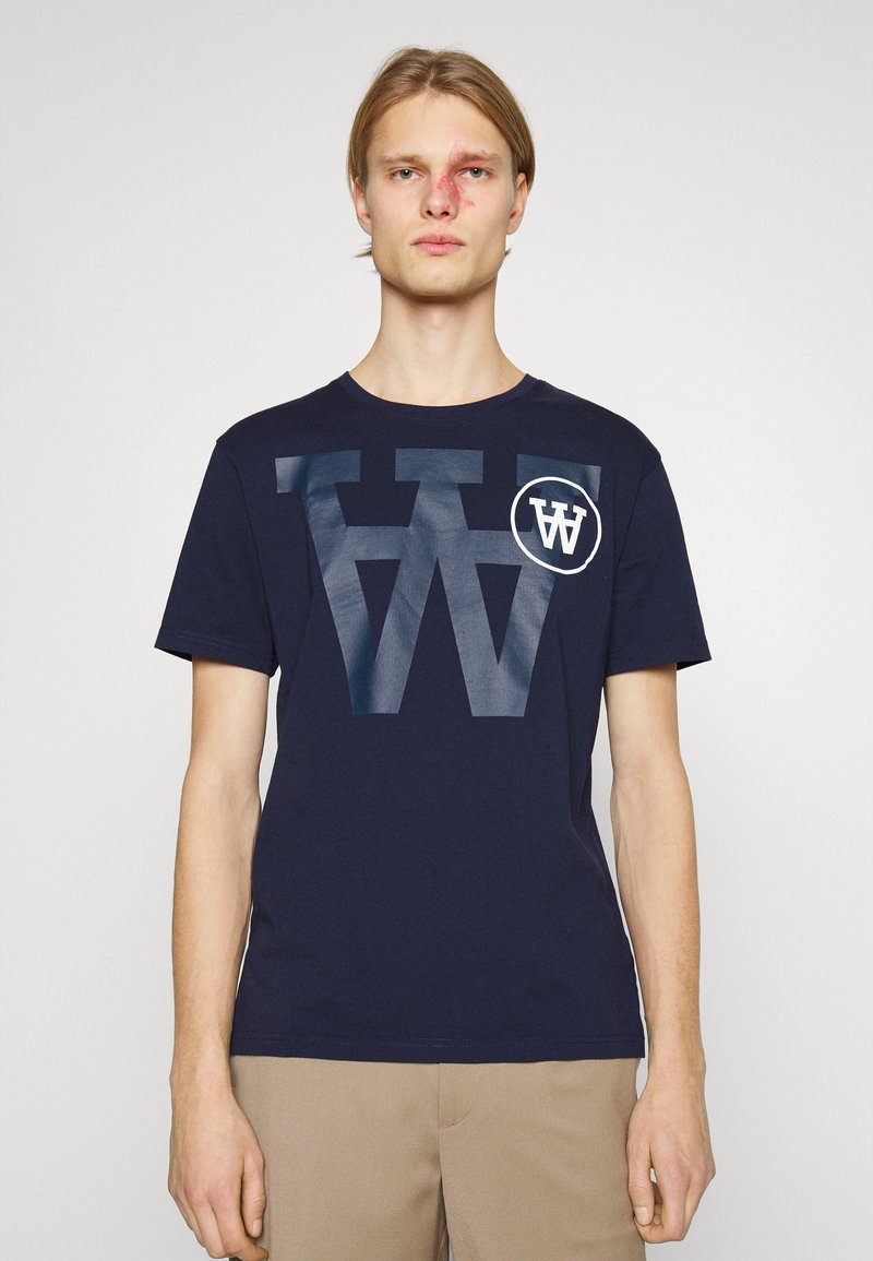 Tricou din bumbac de culoare bleu marin, cu un mare logo alb „W” uzat și un logo accent circular alb pe piept. Mâneci scurte și decolteu rotund.