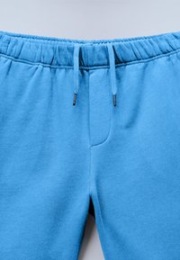 Blauwe katoenen sweatpants met elastische tailleband en aantrekkoorden, gezien van de voorkant van de taille tot het bovenbeen.