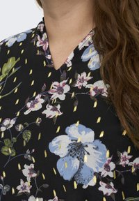 Svart blommig blus med V-ringning, prydd med färgglada blommor och gyllene accenter som är utspridda på tyget, vilket visar ett strukturerat mönster.