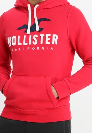 Osoba nosząca czerwoną bluzę z kapturem Hollister California z białymi sznurkami i rękami w przedniej kieszeni, pokazana od klatki piersiowej do bioder.