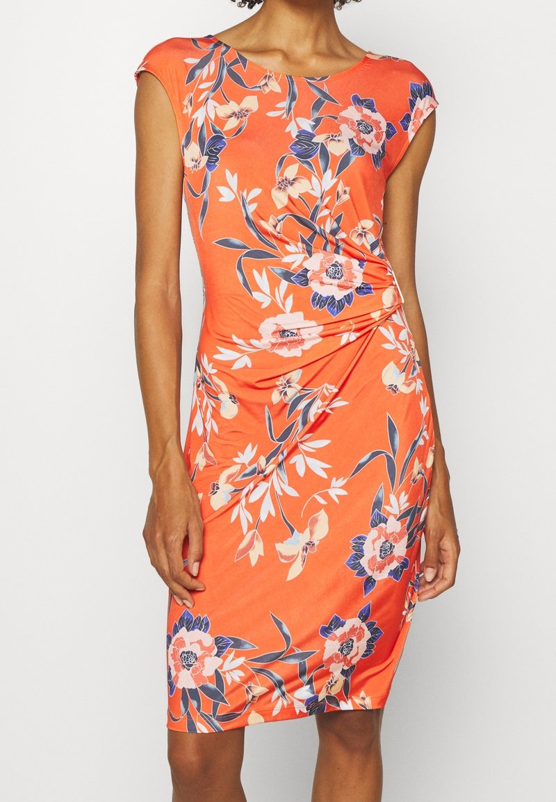 Femme portant une robe orange sans manches, longueur genoux, avec des motifs floraux roses et bleus et un tissu froncé à la taille.