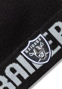 Mustakarvainen pipo, jossa on harmaa "Raiders"-logo-patch ja brodeerattu kilpi hopea-aksonteilla, esittelee teksturoitua kangasta.