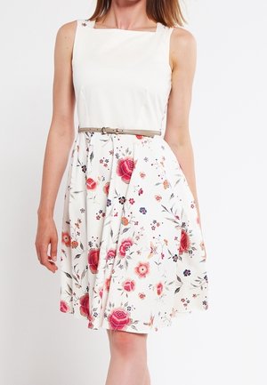 Vrouw draagt een mouwloze witte jurk met bloemenrok en dunne beige riem, staand tegen een effen witte achtergrond.