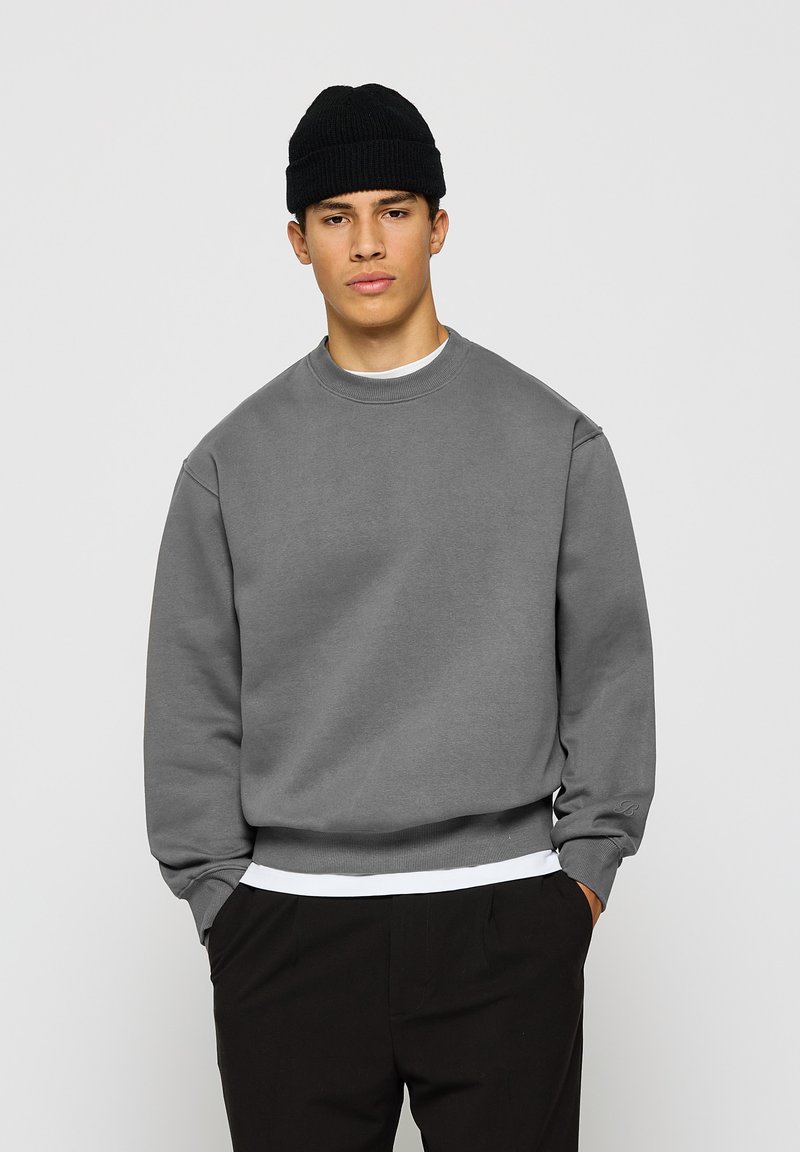 Ung mann med svart lue, grå sweatshirt over hvit skjorte, og svarte bukser med hender i lommene mot hvit bakgrunn.