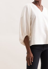 Hvid, struktureret bluse med V-udskæring og voluminøse ærmer samlet ved manchetterne. Samlet med mørke jeans.