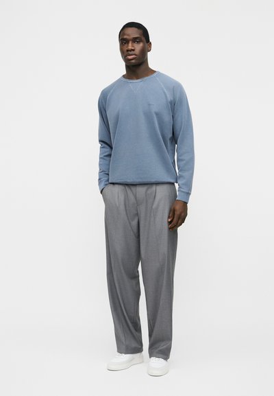 Homme debout portant un sweat-shirt à manches longues bleu, un pantalon large gris et des baskets blanches, les mains dans les poches, sur un fond uni.