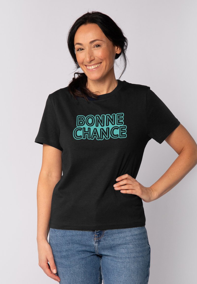 Schwarzes Kurzarm-T-Shirt mit "BONNE CHANCE" in türkisfarbiger Schrift. Lässiges Design mit glatter Textur, kombiniert mit blauen Jeans.