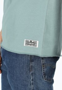 T-shirt azzurro chiaro con maniche corte, orlo curvo e un'etichetta tessuta che recita "DENIM FACTORY" in basso. Indossato con jeans blu.