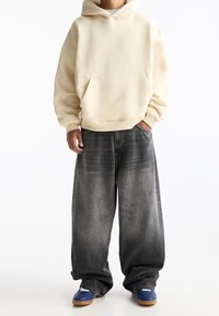 Béžová oversized mikina s přední kapsou; kombinována s širokými černými denimovými džíny s gradientním efektem. Modré boty dokončují vzhled.
