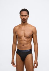 Μαύρες βαμβακερές εσώρουχες με ελαστική ζώνη που φέρει λογότυπο. Το ύφασμα είναι λείο με εφαρμοστή σχεδίαση, τονίζοντας τις καμπύλες του σώματος.