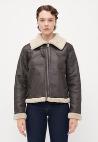 Kvinne iført en mørkebrun shearling-jakke med kremfarget pelskrage, mansjetter og nederkant, kombinert med mørkeblå jeans, vendt mot oss.