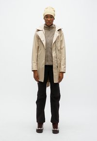 Μπεζ ελαφρύ αδιάβροχο μπουφάν με κουκούλα, φερμουάρ στο μπροστινό μέρος, γκρι εσωτερικό στρώμα από φλις, μαύρο παντελόνι και καφέ πεζοπορικά μποτάκια.