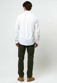 Man staat met zijn rug naar ons toe, gekleed in een wit longsleeve shirt, donkergroene broek en beige laarzen op een effen lichte achtergrond.