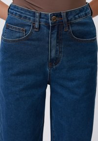 Mėlyni plačių kojų džinsai su aukštu liemeniu, su oranžinėmis siūlėmis, mygtuko uždarymu ir klasikinio penkių kišenių dizainu.