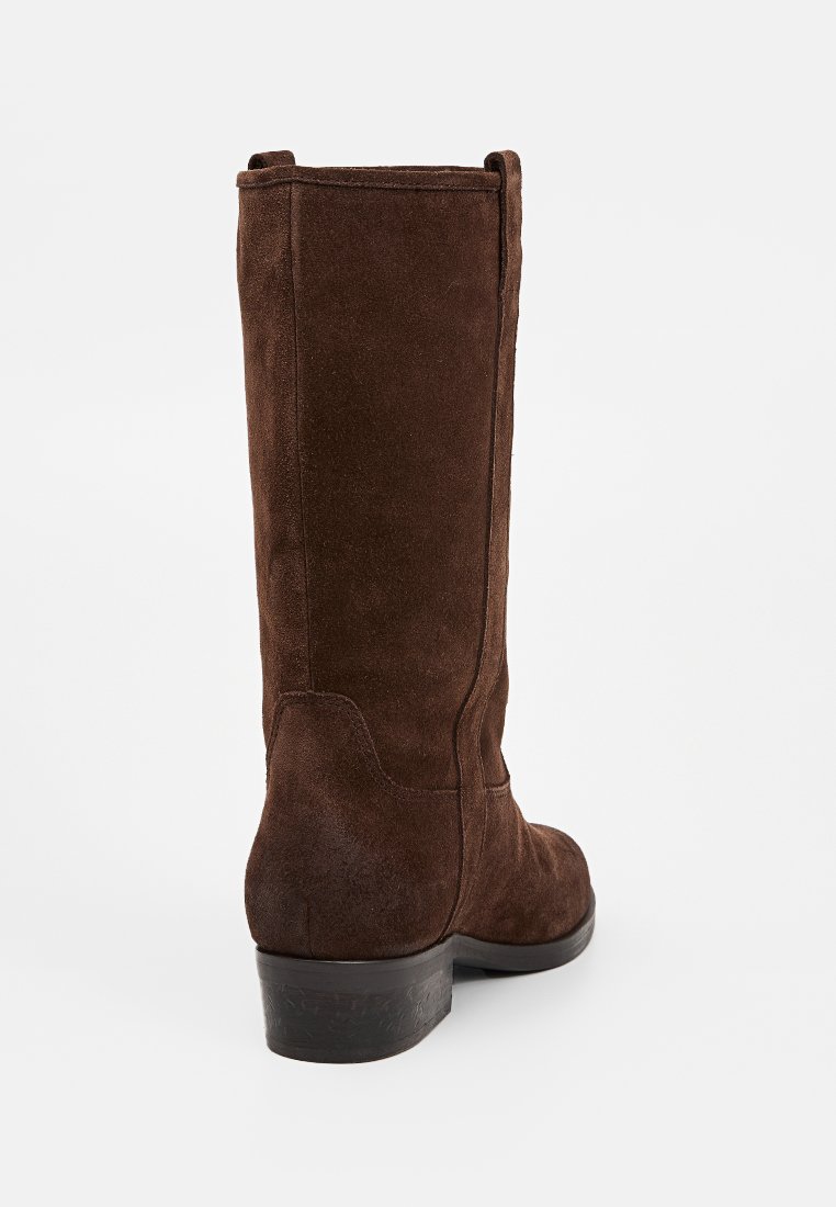 Stivale marrone in suede alto fino al ginocchio, con punta rotonda, texture liscia e tacco basso incassato. Presenta un delicato dettaglio di cucitura lungo i lati.