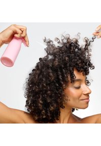 Un flacon pulvérisateur avec un corps en plastique rose, utilisé pour vaporiser les cheveux ; le modèle a des cheveux bouclés foncés et volumineux avec des reflets plus clairs et les yeux fermés.