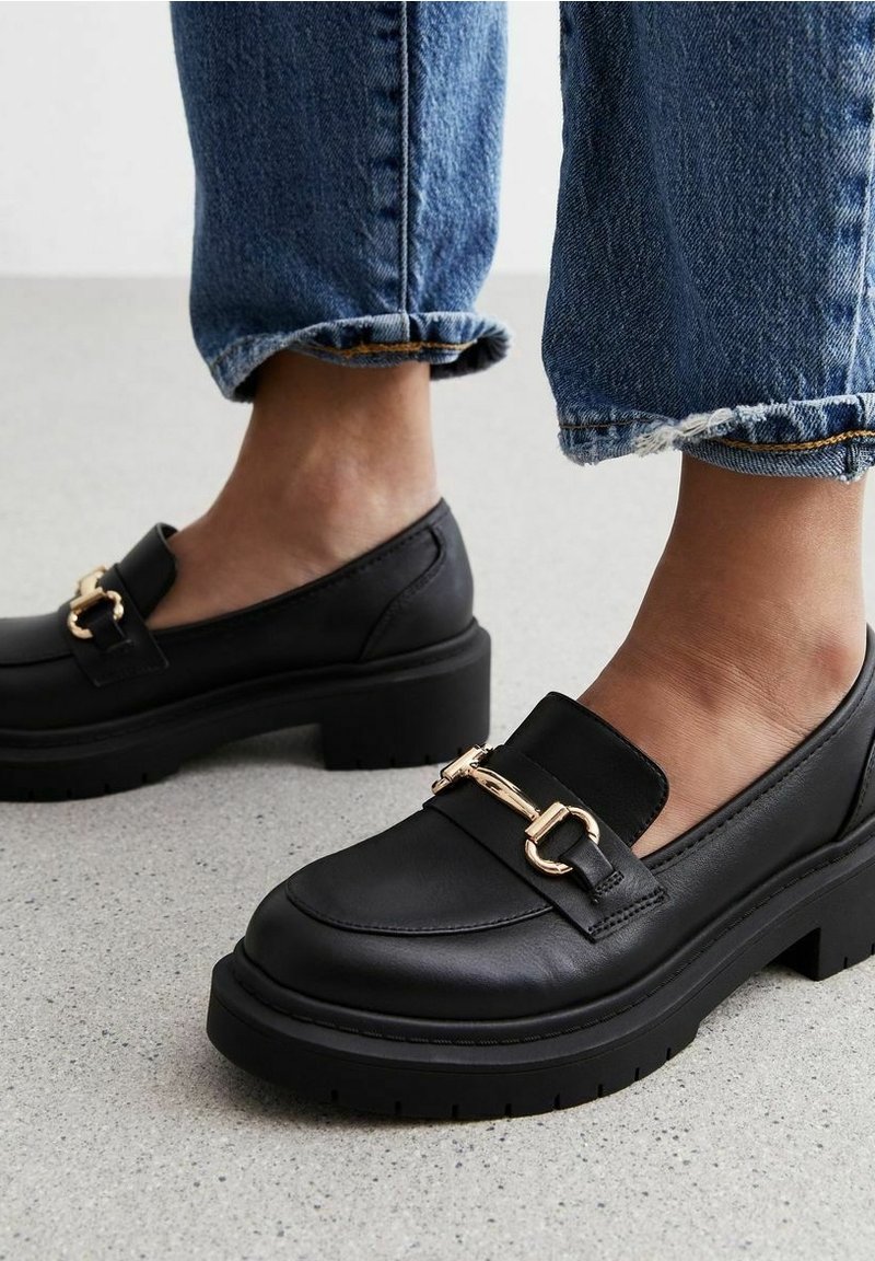 Des mocassins noirs en cuir avec plateforme, dotés d'une semelle épaisse, d'un bout rond et d'un accent décoratif en chaîne dorée à l'avant.