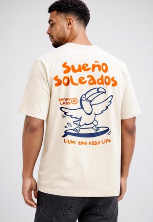 Man wearing a cream t-shirt with orange and blue graphic of a toucan surfing and text "Sueño Soleados" and "Livin' the easy life" on the back.