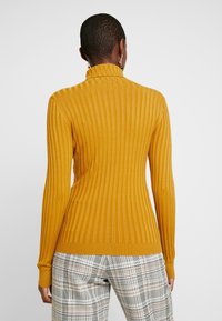 Senapsgul ribbad polokragetröja med långa ärmar och figurnära passform, i kombination med rutig byxa i flera färger.