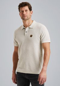 Hellbeige Polo-Shirt aus strukturiertem Stoff, mit einem Kragen, zwei Knöpfen und einem kleinen Logopatch auf der Brust.