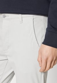 Pantaloni din bumbac gri deschis, cu o croială slim, având un buzunar frontal și închidere cu nasture. Talie este parțial acoperită de un top bleumarin.